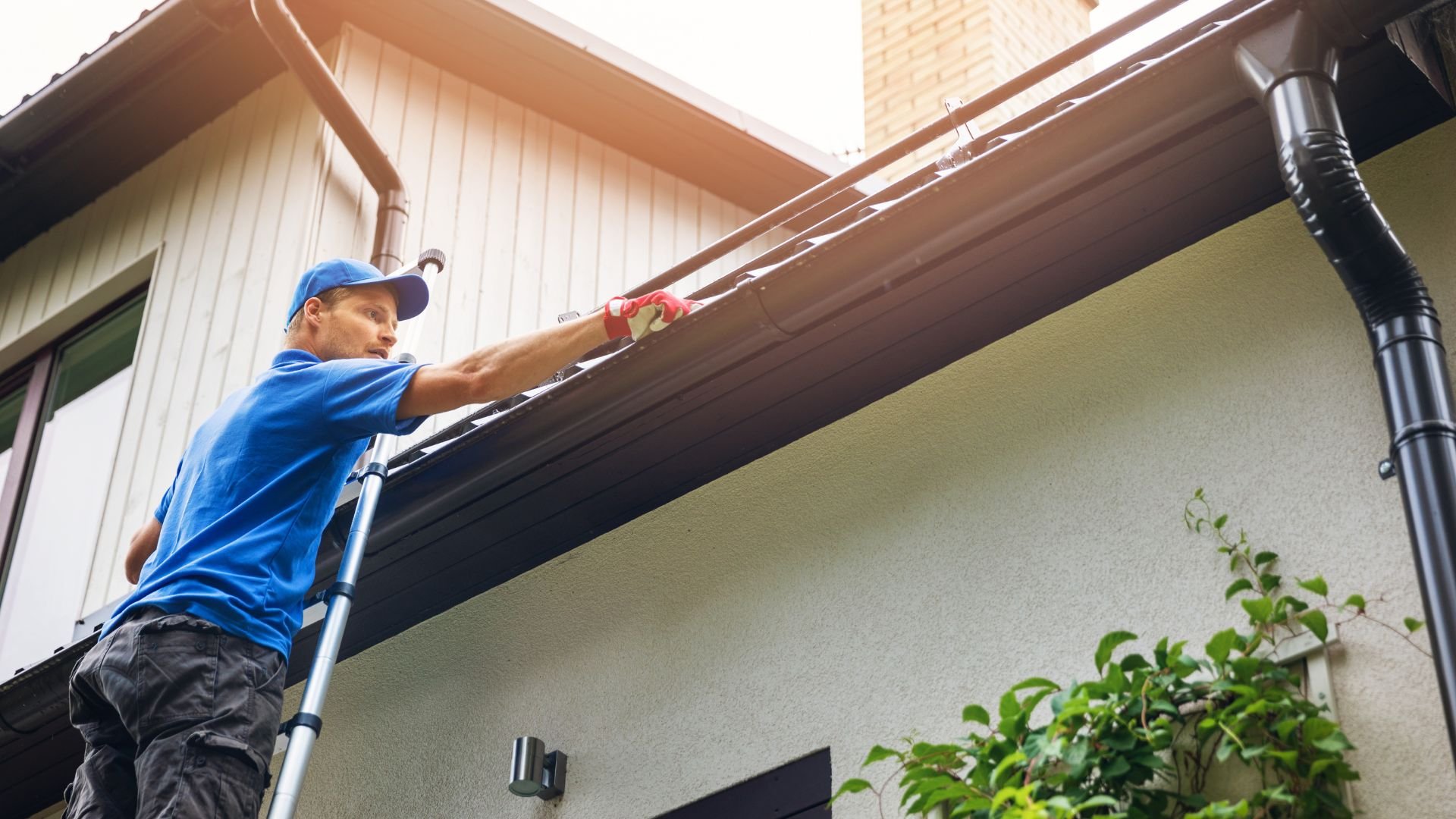 Worker on ladder cleaning gutters of residential house on sunny day